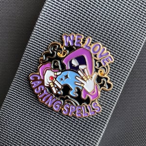 May include: A gold-colored enamel pin with a purple-clad figure casting a spell. The figure is holding a blue orb with a white star. The text around the figure reads "We Love Casting Spells".