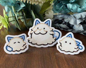 May include: Three stickers featuring a cartoon cat design. The stickers are white with light brown and blue accents. The cat has a fluffy appearance and a smiling expression. The stickers are arranged on a dark brown wooden surface.