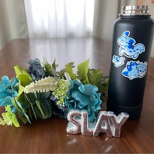 May include: A black stainless steel water bottle with three blue and white cartoon animal stickers. The bottle is sitting on a wooden table with a silver metal sign that says "SLAY" and a bouquet of blue and green flowers.