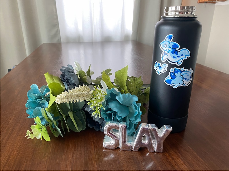 May include: A black stainless steel water bottle with three blue and white cartoon animal stickers. The bottle is sitting on a wooden table with a silver metal sign that says "SLAY" and a bouquet of blue and green flowers.