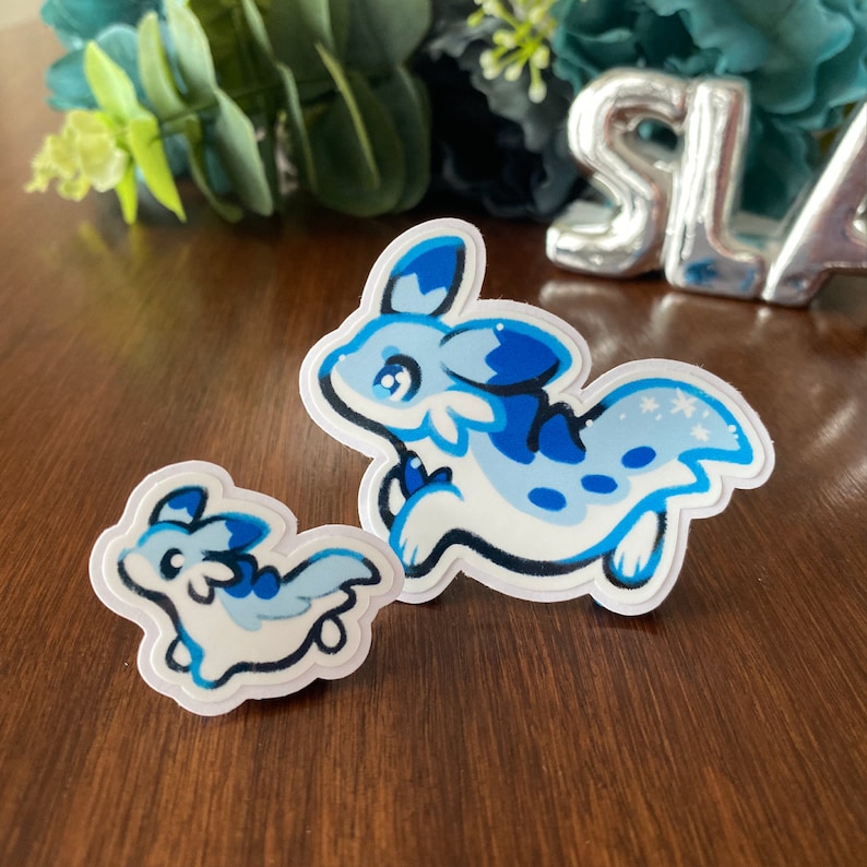 May include: Two blue and white cartoon animal stickers. The larger sticker features a blue and white animal with a long tail and a blue fin. The smaller sticker features a smaller version of the same animal.