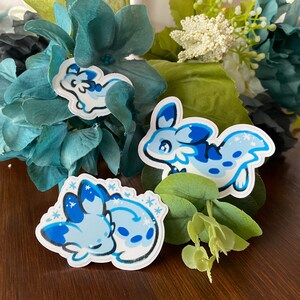 May include: Three blue and white sea slug stickers with snowflake designs. The stickers are on a wooden surface with green foliage.