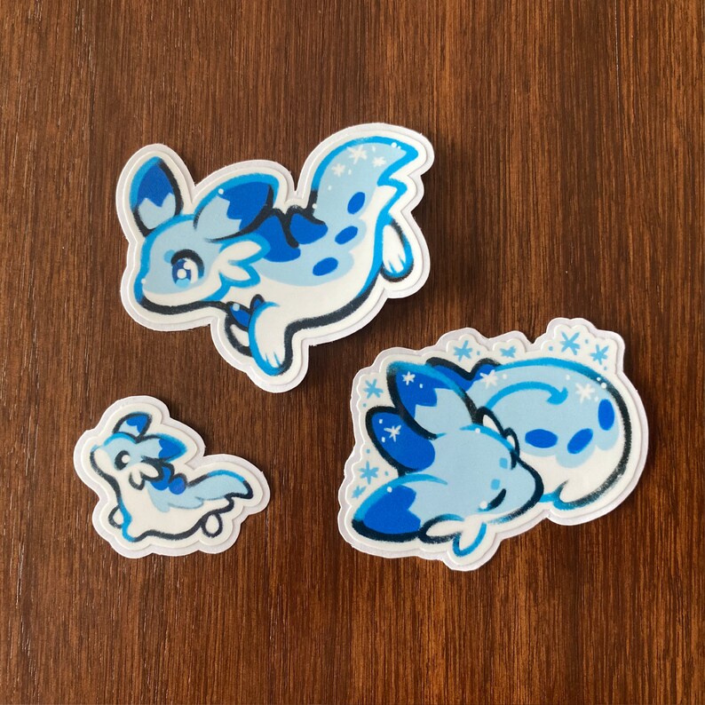 May include: Three blue and white cartoon sea slug stickers with snowflake designs. The stickers are on a brown wooden surface.