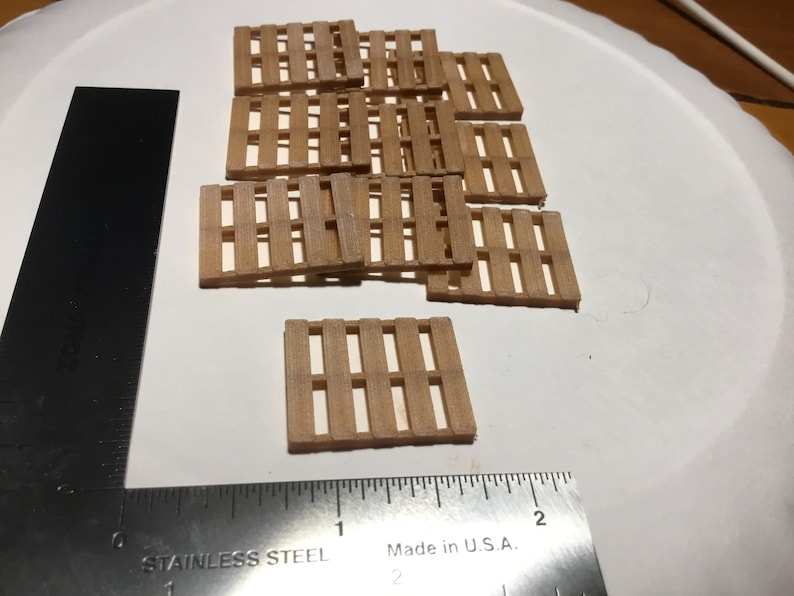 May include: A collection of small, light brown wooden pallets arranged on a white surface. A stainless steel ruler is positioned to the left, with the text "Made in U.S.A." visible. The pallets are miniature, likely for crafts or model building.