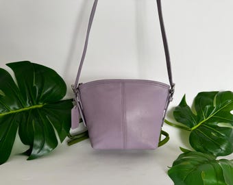 May include: A lavender leather crossbody bag with a curved top and a long, thin strap. The bag has a small tag attached and silver-tone hardware. The background includes large green leaves.