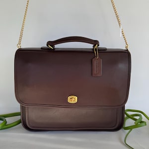 May include: A dark brown leather briefcase featuring a gold-tone turn-lock closure, a top handle, and a detachable gold-tone chain shoulder strap. A matching leather luggage tag embossed with the word COACH hangs from the handle.