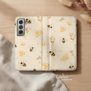 May include: A beige phone case with a bee and honeycomb pattern. The case is open, revealing the phone's camera. The design includes yellow bees, honeycombs, and floral accents. The case is made of a textured material.