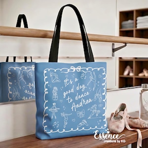 May include: A light blue tote bag with black handles, featuring white line art of ballet dancers and the text "It's a good day to dance Andrea". The bag is on a wooden surface with ballet shoes and a water bottle.