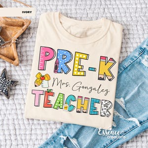 May include: Ivory-colored t-shirt with a colorful graphic that reads "PRE-K Mrs. Gonzalez TEACHER." The design includes a ruler, crayon, pencil, and apple. The shirt is folded on top of a pair of blue jeans.