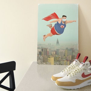 Puede incluir: Un par de zapatillas Nike Air Max 1 blancas y beige con detalles rojos y un logotipo Swoosh rojo. Las zapatillas tienen una entresuela blanca y una suela exterior beige. Las zapatillas están sobre una superficie blanca delante de un póster de un hombre volando con una capa roja.