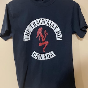 Op de afbeelding: Zwart T-shirt met een rode afbeelding van een gevleugelde figuur die een schedel vasthoudt, met de tekst "THE TRAGICALLY HIP" in een witte boog erboven en "CANADA" in een witte banner eronder. Het shirt hangt aan een zwarte hanger.