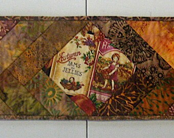 May include: A long, rectangular quilted table runner with a patchwork design. The runner features a variety of warm colours, including gold, brown, and orange, arranged in a diagonal pattern. A central label reads "Jams & Jellies".