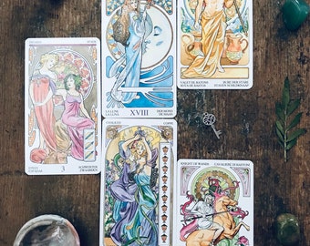 May include: A collection of tarot cards with Art Nouveau illustrations, featuring figures and symbols. The cards are arranged on a rustic wooden surface, accompanied by crystals, a key, and flowers. The cards include text in multiple languages.