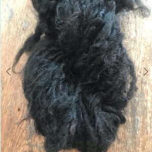 May include: A dense, unspun mass of raw black wool fiber. The material features a highly textured, curly, and tangled appearance with individual strands of dark, coarse hair visible throughout the clump, showcasing a natural, unprocessed state.