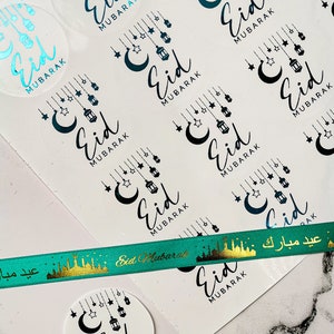 Op de afbeelding: Transparante ronde stickers met de tekst "Eid Mubarak" in zwart script, versierd met manen, sterren en lantaarns. Een groen lint met gouden tekst en een gouden skyline-ontwerp is ook zichtbaar. Voor het islamitische feest Eid.