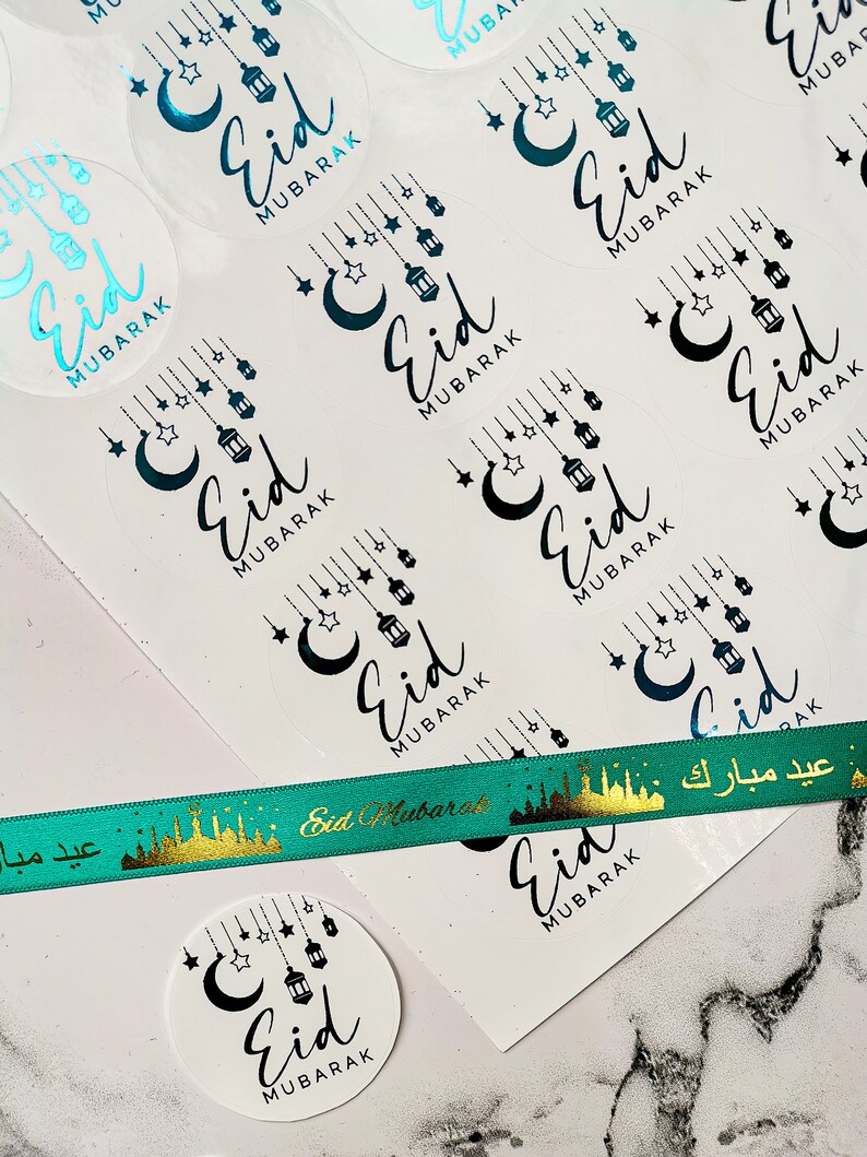 Op de afbeelding: Transparante ronde stickers met de tekst "Eid Mubarak" in zwart script, versierd met manen, sterren en lantaarns. Een groen lint met gouden tekst en een gouden skyline-ontwerp is ook zichtbaar. Voor het islamitische feest Eid.