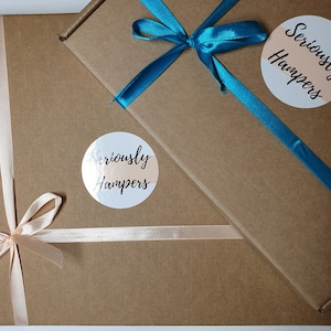 Op de afbeelding: Twee bruine kartonnen geschenkdozen, de ene iets over de andere. De bovenste doos is vastgebonden met een felblauwgroen lint, de onderste met een lichtroze lint. Elke doos heeft een ronde sticker met de tekst "Seriously Hampers".