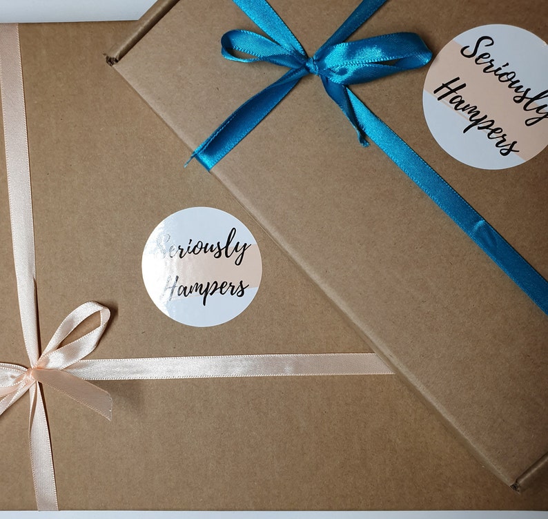 Op de afbeelding: Twee bruine kartonnen geschenkdozen, de ene iets over de andere. De bovenste doos is vastgebonden met een felblauwgroen lint, de onderste met een lichtroze lint. Elke doos heeft een ronde sticker met de tekst "Seriously Hampers".