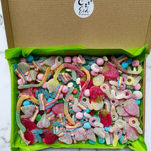 Op de afbeelding: Een kartonnen doos gevuld met een verscheidenheid aan kleurrijke snoepjes, waaronder gummy worms, fruitvormen en gesuikerde lekkernijen. De doos is gevoerd met groen vloeipapier en heeft een ronde sticker met de tekst "Eid Mubarak".