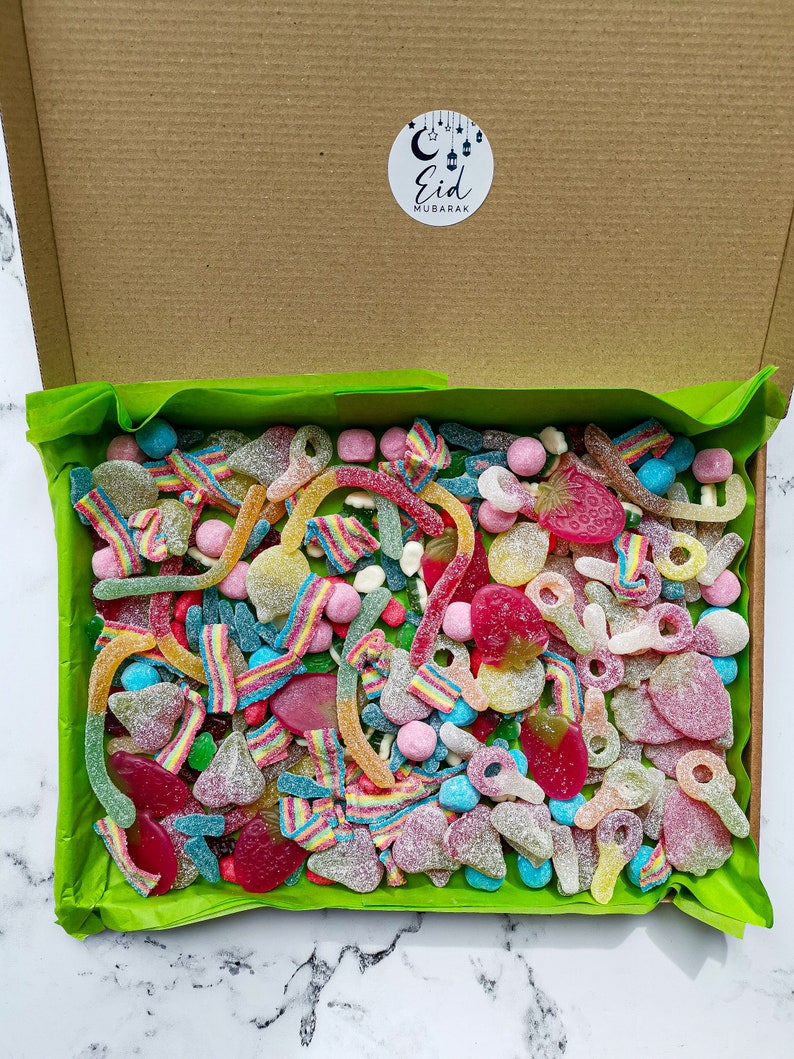 Op de afbeelding: Een kartonnen doos gevuld met een verscheidenheid aan kleurrijke snoepjes, waaronder gummy worms, fruitvormen en gesuikerde lekkernijen. De doos is gevoerd met groen vloeipapier en heeft een ronde sticker met de tekst "Eid Mubarak".