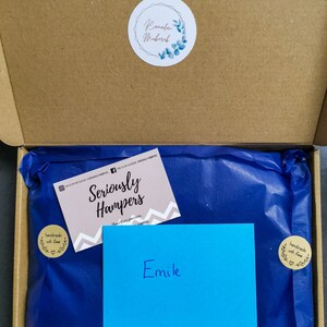 Op de afbeelding: Een open kartonnen doos met een cadeau verpakt in blauw vloeipapier. Een visitekaartje met de tekst "Seriously Hampers" en een blauwe kaart met de naam "Emile" zijn zichtbaar. Een ronde sticker met de tekst "Ramadan Mubarak" staat op de doos.