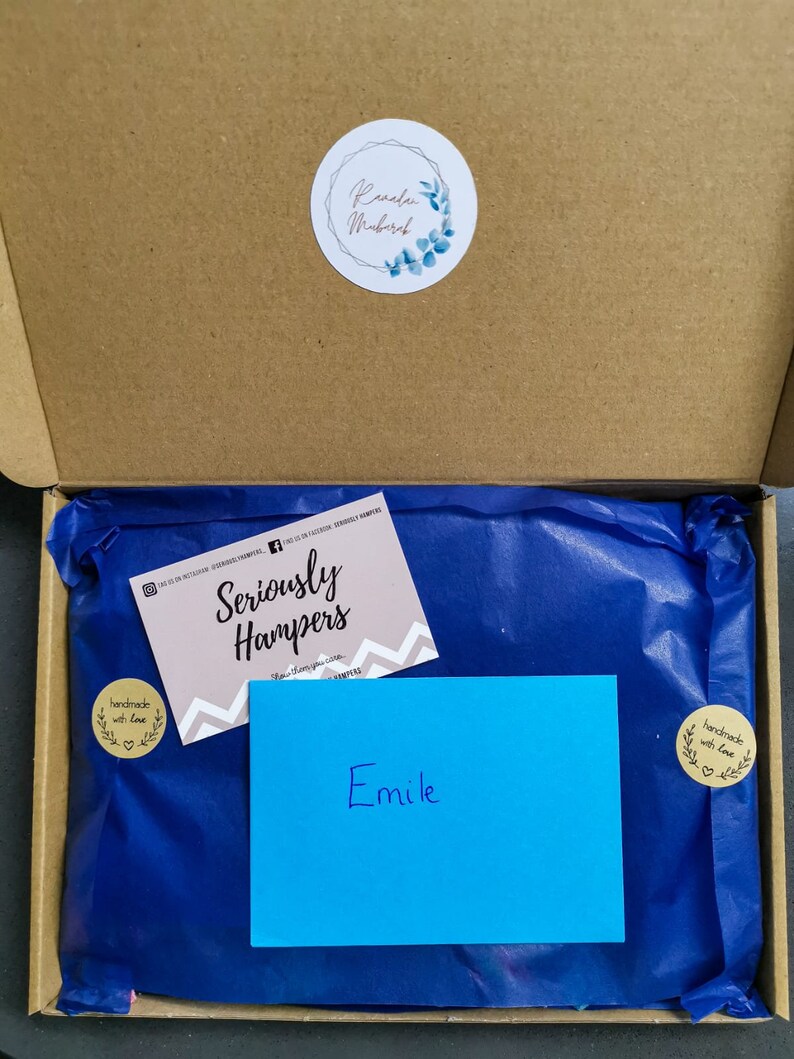 Op de afbeelding: Een open kartonnen doos met een cadeau verpakt in blauw vloeipapier. Een visitekaartje met de tekst "Seriously Hampers" en een blauwe kaart met de naam "Emile" zijn zichtbaar. Een ronde sticker met de tekst "Ramadan Mubarak" staat op de doos.