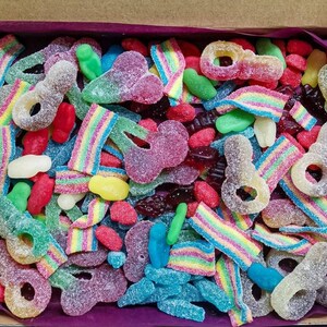 Op de afbeelding: Een doos gevuld met een verscheidenheid aan kleurrijke gummy snoepjes. De selectie omvat regenboogriemen, sleutelvormige gummies en andere vormen in tinten rood, groen, blauw en geel. De snoepjes zijn bedekt met suiker.
