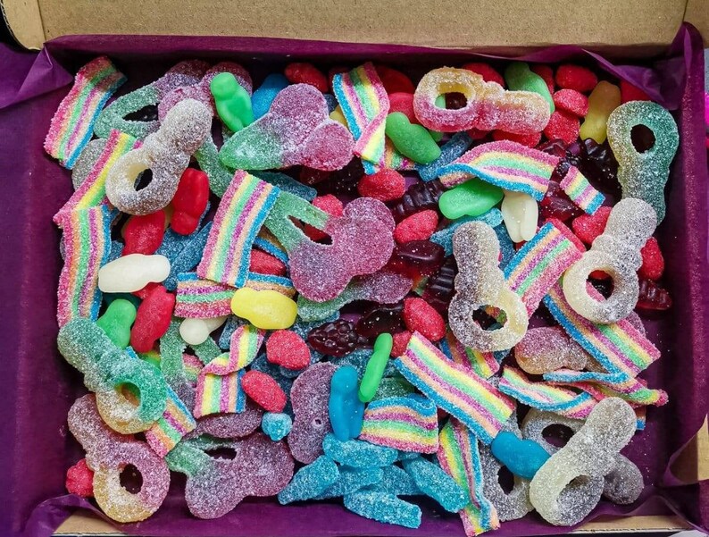 Op de afbeelding: Een doos gevuld met een verscheidenheid aan kleurrijke gummy snoepjes. De selectie omvat regenboogriemen, sleutelvormige gummies en andere vormen in tinten rood, groen, blauw en geel. De snoepjes zijn bedekt met suiker.