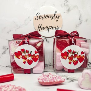 May include: Two clear square gift boxes filled with red and pink sweets and marshmallows, tied with burgundy ribbons. Each box has a round sticker that says "Happy Valentine's Day" with red hearts. The background is a marble pattern.