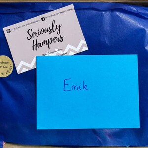 Op de afbeelding: Een geschenkdoos met blauw vloeipapier, een roze visitekaartje en een blauwe kaart met de naam "Emile" erop geschreven. Het visitekaartje bevat de tekst "Seriously Hampers" en social media-handles. Twee "handmade with love" stickers zijn ook zichtbaar.