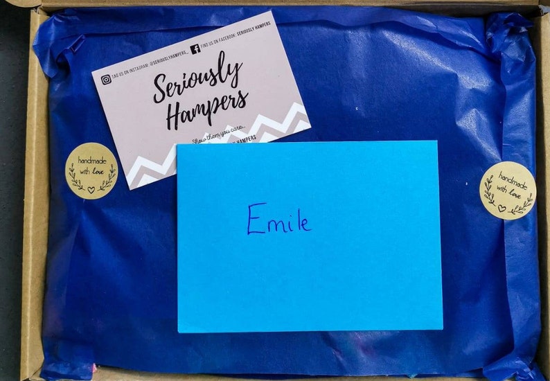 Op de afbeelding: Een geschenkdoos met blauw vloeipapier, een roze visitekaartje en een blauwe kaart met de naam "Emile" erop geschreven. Het visitekaartje bevat de tekst "Seriously Hampers" en social media-handles. Twee "handmade with love" stickers zijn ook zichtbaar.