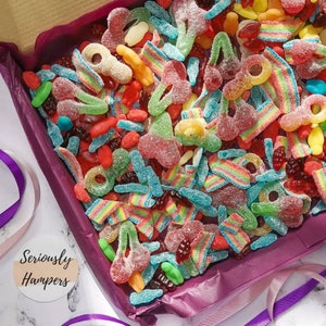 Op de afbeelding: Een doos vol kleurrijke gummy snoepjes. De snoepjes bevatten regenboogriemen, gummy kersen en andere vormen in rood, groen, geel en blauw. De doos is gevoerd met paars papier en heeft een wit en paars lint.
