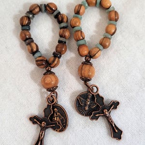 May include: Two wooden rosary rings with olive wood beads and small blue-green beads. Each ring has a decorative copper-colored pendant with a cross and religious imagery. The rosaries are displayed on a white surface.