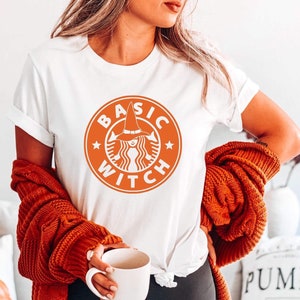 May include: A white t-shirt with an orange graphic of a witch wearing a hat and the text "BASIC WITCH" in a circle.