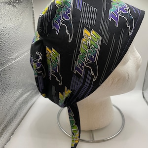 May include: Black surgical scrub hat with a purple and green print of the Tampa Bay Buccaneers logo and the words "Tampa Bay" and "Bucs".