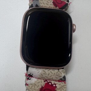 Może przedstawiać: Smartwatch z kopertą z metalu w kolorze różowego złota i czarnym ekranem, wyposażony w beżowy pasek z tkaniny typu scrunchie z powtarzającym się wzorem małych postaci przypominających kreskówki oraz geometrycznych kształtów w kolorze czerwonym, czarnym i białym.