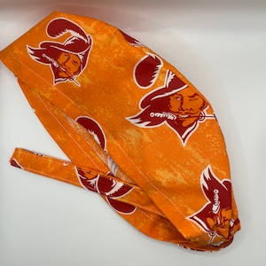 May include: Orange surgical scrub hat with a repeated pattern of a red and white pirate logo. The hat has a tie on the back.