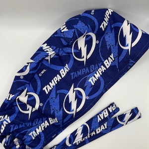 May include: Blue and white surgical scrub hat with a repeating pattern of the Tampa Bay Lightning logo and the words "Tampa Bay".
