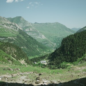 May include: A view of a valley with lush green vegetation and a small building in the distance. The valley is surrounded by mountains, and the sky is blue with some clouds.