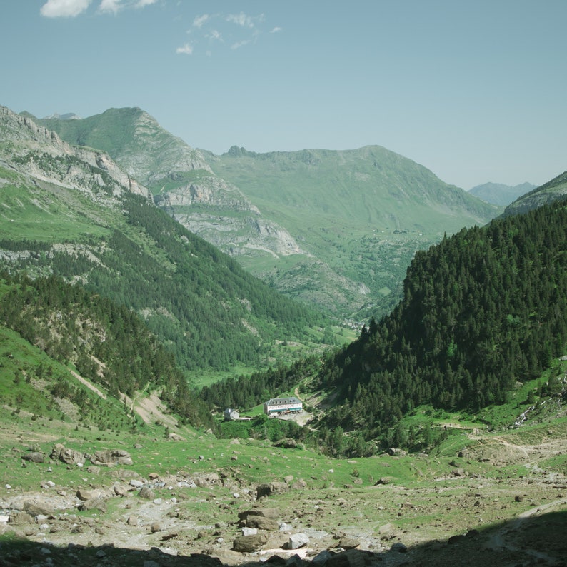 May include: A view of a valley with lush green vegetation and a small building in the distance. The valley is surrounded by mountains, and the sky is blue with some clouds.