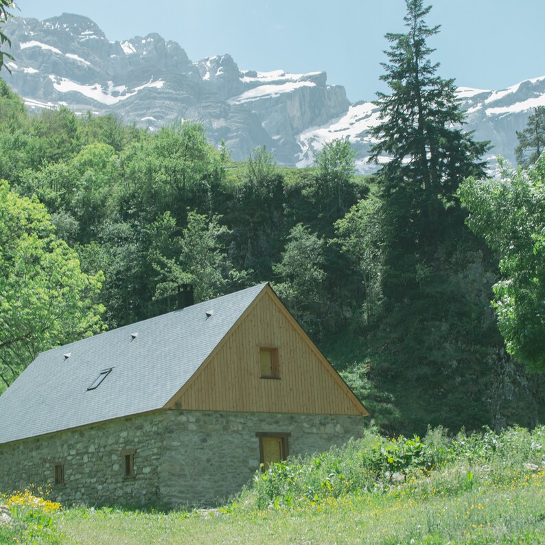 May include: A stone building with a wooden roof and a wooden door. The building is surrounded by green trees and a mountain range in the background.