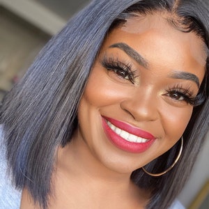 May include: A woman with dark gray hair styled in a bob, wearing gold hoop earrings and red lipstick. She is smiling and looking at the camera.