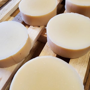 May include: Several round, solid, off-white bars of soap are arranged in a staggered pattern. Each bar has a smooth, flat top surface and straight, vertical sides, showcasing a uniform, pale cream color throughout the entire collection of items.