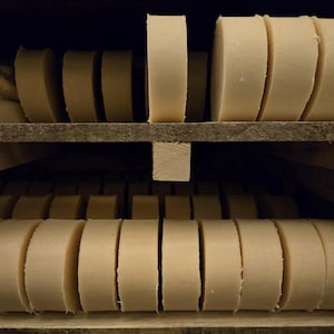 May include: Rows of round, light-colored soap bars are neatly arranged on wooden shelves. The soap bars are cut into thick, circular shapes, with a slightly rough texture. The shelves are made of unfinished wood, creating a rustic, natural aesthetic.