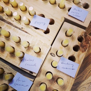 May include: Wooden display with rows of small yellow lip balm tubes. Each row has a blue sticky note with a different flavour written on it, including Eggnog, Salted Caramel, Anise Spearmint, Strawberry Cheesecake, Bourbon Vanilla Coffee.