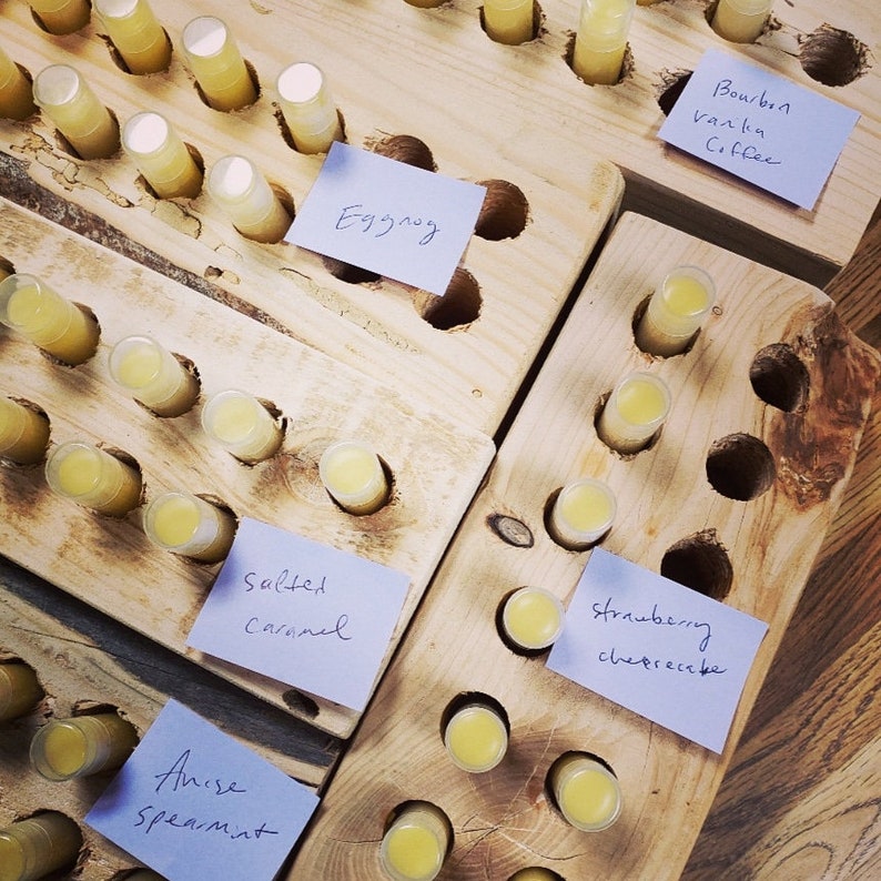 May include: Wooden display with rows of small yellow lip balm tubes. Each row has a blue sticky note with a different flavour written on it, including Eggnog, Salted Caramel, Anise Spearmint, Strawberry Cheesecake, Bourbon Vanilla Coffee.