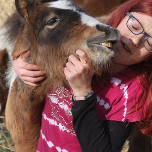 May include: A woman with red hair is hugging a brown and white pony. She is wearing a pink and white tie-dye shirt with black sleeves and a watch.
