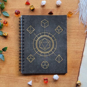 May include: A black spiral bound notebook with a gold design featuring a large 20-sided die and smaller dice around it. The notebook is surrounded by dice on a wooden surface.