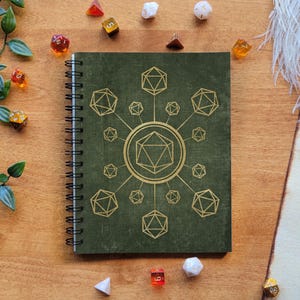 May include: A green spiral bound notebook with a gold design of a 20-sided die in the centre surrounded by smaller dice. The notebook is on a wooden surface with dice scattered around it.