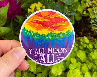 Peut inclure: Un autocollant rond avec un paysage de montagne arc-en-ciel et un soleil dans le ciel. Le texte "Y'all Means All" est écrit en blanc sur un fond violet.